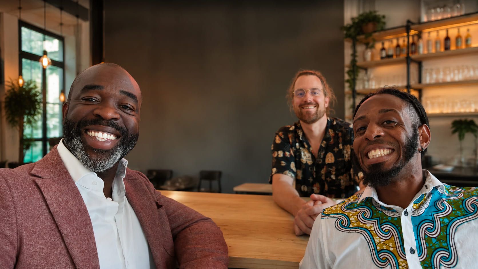 Drie mannen lachen naar de camera aan de bar van een café; twee zitten vooraan, de derde staat erachter